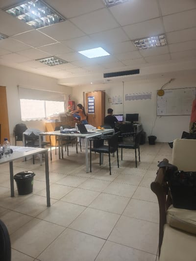 A brightly lit office space features fluorescent ceiling panels and tiled flooring. Two people are present: one near a window amidst boxes, and another seated at a computer desk. Several tables and chairs are scattered throughout the room, some with laptops on them. A whiteboard is visible on the right wall, along with other wall-mounted notices. There's a trash bin near a table in the foreground, and a couch with a handbag resting on it.