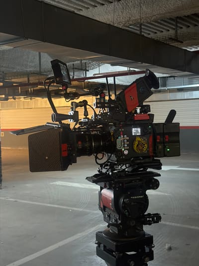 The image displays a professional cinema camera rig, fully assembled and in what appears to be an industrial or urban indoor setting, possibly a parking garage or warehouse, in San Sebastián De Los Reyes, Spain. The camera is positioned upright, equipped with various accessories for a production shoot.
The main subject is a high-end cinema camera body, possibly an ARRI, fitted with a prominent ZEISS lens, whose brand name is clearly visible. Attached to the front of the lens is a matte box, and a follow focus system encircles the lens barrel. An external monitor or electronic viewfinder is mounted to the side of the rig, displaying camera settings. A wireless video transmitter/receiver, featuring a red and black design, is also integrated into the setup. The camera appears to be mounted on an OConnor fluid head, with "oconnor" visible on its side. The entire rig is supported by a system of rods and mounting hardware, indicating a complex and robust setup.
A distinctive yellow sticker or patch is affixed to the side of the camera body, depicting a cartoonish character, possibly a duck or bird in a chef's hat. Partially visible text on the sticker includes "THE 7" and "BIG". A small display screen on the camera body shows icons and numerical values, likely indicating camera parameters such as resolution, frame rate, or battery status.
The background consists of concrete structures, overhead ducts or pipes, and what appear to be faint markings on the ground. The lighting suggests an indoor or nighttime setting, with artificial illumination creating a functional but dim atmosphere. There are no people visible, and the scene is focused entirely on the technical equipment, indicating the camera is either awaiting action or on standby during a film production.