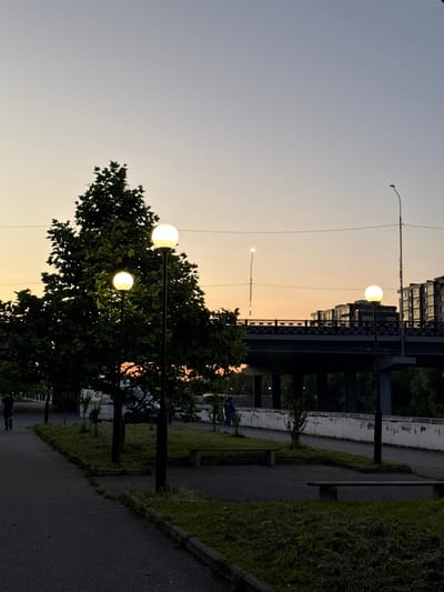 The image captures an outdoor scene at dusk, possibly in Kamysh-Gadzhiaul, Russia. The sky displays a gradient of light blues and pale yellows, suggesting the sun has recently set. Several streetlights with glowing globes are visible, some illuminating nearby trees and a paved pathway. One streetlight in the foreground is particularly prominent, with a large, illuminated sphere. Beyond the pathway, a line of grassy embankment leads to a building with multiple windows, and a dark structure resembling a bridge or overpass stretches across the middle ground. The overall atmosphere is tranquil, with the warm glow of the streetlights contrasting with the deepening twilight. The presence of a pathway and benches hints at a public park or promenade. A few indistinct figures can be seen on the pathway in the distance, suggesting people are present, perhaps enjoying the evening. The vegetation is lush and green, indicating a temperate climate. There is no visible text in the image.