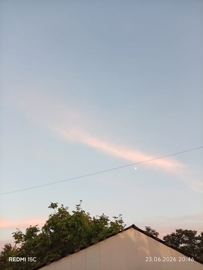 The image captures a tranquil evening sky over Mykolaivka, Ukraine. The sky is a soft, pale blue, transitioning to lighter hues near the horizon, indicative of dusk. A delicate, elongated cloud, tinged with a faint pink from the setting sun, stretches diagonally across the upper half of the frame. A sliver of the moon is visible just below the cloud, a subtle white crescent against the blue.
In the foreground, the sharp angle of a light-colored roof cuts across the bottom of the image. Lush green foliage from trees peeks over the roofline, adding a touch of life and natural beauty to the scene. A thin, dark wire runs horizontally across the sky, a man-made element contrasting with the natural elements.
The overall mood is peaceful and serene, with the soft light and the quiet presence of the moon and clouds suggesting the end of a day. The timestamp in the bottom right corner, "23.06.2026 20:46," confirms the time of day. The text "REDMI 15C" in the bottom left corner indicates the device used to capture the photograph. There are no people or specific activities depicted, leaving the focus on the serene natural landscape and the quiet beauty of the evening sky.