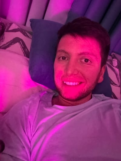 A man is lying down on a bed, with pink and purple lighting illuminating the scene. He is looking directly at the camera with a smile. Behind him, there are pillows and a headboard. The man is wearing a light blue t-shirt. The environment suggests a bedroom.