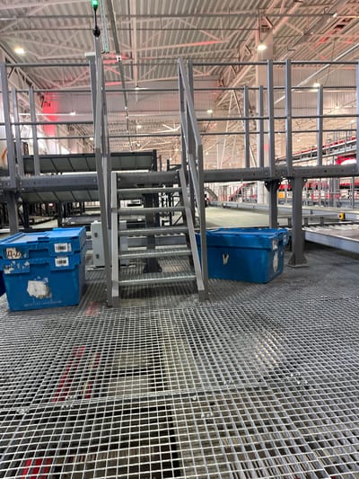 An indoor shot captures a section of a large industrial building, possibly a warehouse or distribution center, in Nerubais'ke, Ukraine. The foreground is dominated by a metal grating floor, suggesting a raised platform. To the left, two blue plastic crates are stacked, one on top of the other, with visible labels and markings. To the right of the crates is a metal staircase with a railing, leading upwards. The background reveals more of the industrial infrastructure, including elevated walkways with railings, metal shelving units, and lighting fixtures on the ceiling. Red lighting is visible in the upper background, hinting at possible operational indicators or aesthetic choices within the facility. The overall environment appears to be clean and organized, typical of a logistics or manufacturing setting. No people are present in the frame.