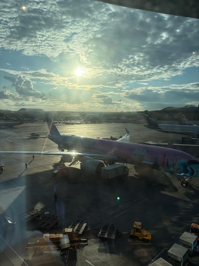 An airport tarmac scene is captured from a high vantage point, possibly from an airplane window. The sun shines brightly through a cloudy sky, casting long shadows across the tarmac. Several airplanes are visible, including a prominently featured aircraft with a colorful livery depicting a cityscape and waves. Ground crew members, dressed in reflective vests, are seen walking on the tarmac. Various airport vehicles, such as baggage carts and a tow tractor, are parked or in motion. In the background, airport buildings and hangars are discernible. The general location is identified as an airport near Искра Снт (чашниково), Russia. The overall atmosphere suggests a busy operational day at the airport.