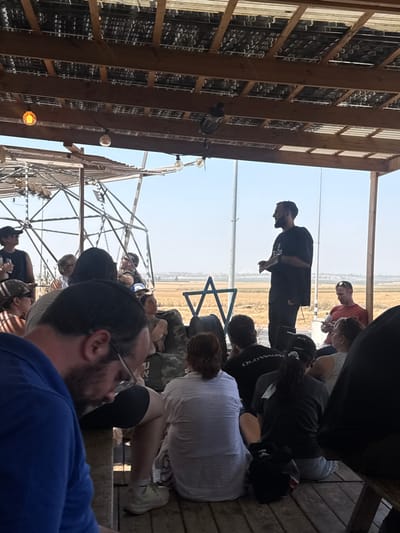 The image depicts an outdoor gathering within the Sdot Negev Regional Council, Israel, under a wooden pergola roof with corrugated panels and embedded glass bottles. A bearded male in a black t-shirt and dark pants stands centrally, gesturing with his right hand as if addressing an audience. Approximately 15-20 individuals, male and female, are seated before him on wooden benches and the wooden platform floor, listening. A man with a beard and glasses, wearing a blue polo shirt and a black kippah, is prominently in the left foreground, seen from behind.
The setting is open-air, with a wide, dry agricultural field stretching into the background under a clear blue sky. A prominent blue metal Star of David sculpture is situated in the field behind the speaker. To the left, a tall metal lattice structure is visible, and a utility pole stands further back. Edison-style light bulbs hang from the pergola's wooden beams. The overall scene suggests a communal educational or informational event taking place in a rural environment.