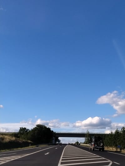 The image depicts a highway scene under a clear blue sky with scattered white clouds. In the foreground, a dark-colored off-road vehicle with a spare tire mounted on the back is driving on the right lane of the asphalt road. The road has clear white lane markings, including directional arrows. In the background, there is a highway overpass and a line of trees and vegetation on both sides of the road. A blue sign with the number "4" is visible on the right side of the road. The scene appears to be in a rural or suburban setting. The location is identified as Předotice, Czechia. There are no people visible in the image.