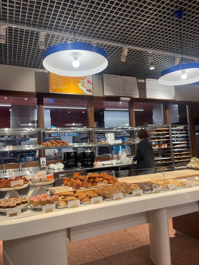 This image depicts the interior of a contemporary bakery or cafe located in Crawley, United Kingdom. The scene is dominated by a brightly lit display counter in the foreground, showcasing a variety of baked goods, with a food preparation area visible through glass partitions in the background. The ceiling features a white grid-patterned system, fitted with numerous square recessed lights and track lighting fixtures. Two prominent circular pendant lights, each with a blue exterior rim and a white interior reflecting a bright bulb, hang centrally over the display area. Behind the main counter, large rectangular windows with dark brown frames offer a view into what appears to be a back-of-house kitchen or preparation area. Reflections on the glass display an inverted word, possibly "INNOVATION" or "INFORMATION." Within this area, a person in a white shirt is partially visible working behind a stainless steel counter equipped with sinks. Stainless steel shelving units are stacked with numerous baked goods, including croissants. Two large, black, lidded urns are positioned on the counter, alongside a wall-mounted hand sanitizer dispenser and stacks of disposable cups. Further to the right, vertical racks laden with baked items are visible. Above the display counter, three menu boards are suspended. The leftmost board, with an orange background, lists "ICED DRINKS" and includes items like "BLACK SESAME ICED MATCHA," "TIRAMISU COLD BREW," "MANGO COLD BREW," "BLACKBERRY COLD BREW," "CHERRY & CHIA ICED TEA," "RASPBERRY & HIBISCUS ICED TEA," "STRAWBERRY & DARJEELING ICED TEA," and "ICED TEA," with prices denoted in British Pounds (£). To its right, a white menu board titled "FROM THE BARISTA" details "HOT DRINKS" and "ICED DRINKS." The third white board, titled "FROM THE KITCHEN," outlines "PORRIDGE," "SOUP & SOURDOUGH," "BRUNCH," and "SALAD POTS." In the midground, behind the main display counter, a person with curly brown hair, dressed in a black jacket and blue jeans, stands facing away from the viewer. This individual appears to be a staff member, possibly attending to the display or serving customers. The foreground is dominated by a long, white, stone-textured display counter with two large, rounded support legs. The counter is laden with an assortment of pastries and cakes presented on various marble stands and wooden boards. Visible items include small tarts or cakes labeled "SEA SALT CARAMEL" and "BANANA & PECAN CAKE," round cakes with purple floral toppings labeled "HONEY CAKE," and square cakes with pink icing identified as