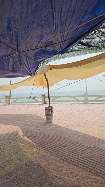 The image, oriented in landscape, captures a sunlit coastal scene in Bac Cam Ranh, Vietnam, featuring various makeshift structures and the ocean in the background.

In the foreground to the left, a large, dark blue tarp or heavy fabric is visible, appearing crumpled and taut in places. It is secured by ropes, some with visible knots, suggesting it might be part of a temporary shelter or covering. Above and behind the blue tarp, a lighter, possibly green or black mesh netting is partially visible.

To the right of the blue tarp and extending towards the center, a light yellow or tan fabric structure, resembling a canopy or sail, billows softly. This fabric is supported by a rough wooden pole or branch, which is anchored into a weathered, cylindrical concrete block or pot resting on the paved ground. Several thin ropes or wires extend from this yellow fabric.

The right side of the image features a paved ground or floor, made of terracotta-colored tiles. The paving displays two distinct patterns: a section with parallel, ribbed lines, and another section with an embossed, possibly floral or geometric, decorative motif. Long shadows are cast across this paved area, indicating bright sunlight, likely during mid-morning or late afternoon.

Along the edge of the paved area, a white concrete railing or fence is visible, featuring vertical posts and angled horizontal bars. Beyond this railing, a tranquil expanse of light blue-green ocean stretches to the horizon under a clear, bright sky. No people or visible text are present in the scene.