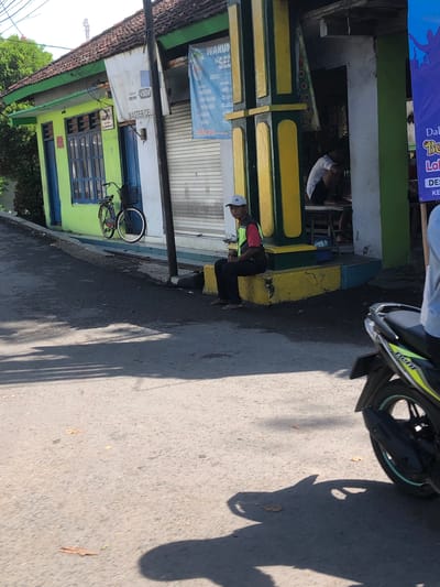 The image shows a street scene in Banjarkemantren, Indonesia, with a building on the right and a street in the foreground. The building appears to be a shop with a green facade and blue doors and windows. A bicycle is parked in front of the shop. A man wearing a red shirt, a yellow vest, black pants, and a white cap is sitting on a yellow concrete ledge in front of the building. A motorcycle is parked on the street in the lower right corner of the image. The ground is asphalt and there are shadows cast by the building and a tree. The sky is visible in the upper portion of the image and appears to be clear.