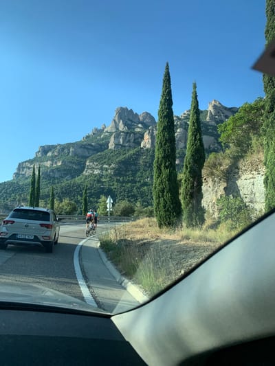 The image shows a scenic mountain landscape, likely Montserrat, near Castellbell i el Vilar, Spain. The foreground is dominated by the interior of a vehicle, with the dashboard and windshield visible. Outside, a paved road curves to the right, marked with a white line. On the road, a silver Volkswagen T-Roc is parked on the left, and two cyclists are riding ahead on the right side of the lane. The road is bordered by dry grass and a steep, rocky incline covered with trees. Towering cypress trees stand on the right side of the road, framing the view of the rugged, green-covered mountains in the background, which are under a clear blue sky. The lighting suggests it is daytime, possibly late afternoon, given the shadows.