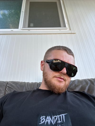 In Shumentsi, Bulgaria, a man with a short haircut and a beard is seated on a dark brown leather sofa, facing the camera. He is wearing a black t-shirt with a white graphic logo that reads "BANDITTT" and appears to be from a brand. The man is also wearing large, dark sunglasses with red trim. Behind him, a white wall with vertical paneling is visible, and above that, a white-framed window is partially open, revealing textured glass on the left and darker glass on the right. The overall scene suggests an outdoor or semi-outdoor setting, possibly a balcony or patio.