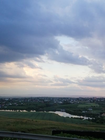 A wide-angle, high-angle shot captures a scenic landscape overlooking a village and a body of water, likely the river that runs through Bulai, Romania. The sky is filled with dramatic, heavy clouds, ranging in color from deep blue to lighter shades of gray and white, with hints of pink and orange near the horizon, suggesting either dawn or dusk.

Below the sky, rolling hills covered in lush green fields stretch towards the distant horizon. In the mid-ground, a settlement is visible, characterized by numerous houses with red and white roofs, nestled amongst trees. A winding river or lake reflects the sky, its surface calm. The foreground features a grassy slope with a weathered metal guardrail running horizontally across the bottom of the frame. The overall impression is one of a tranquil, rural setting at the edge of a developed area. There are no people visible in the image.