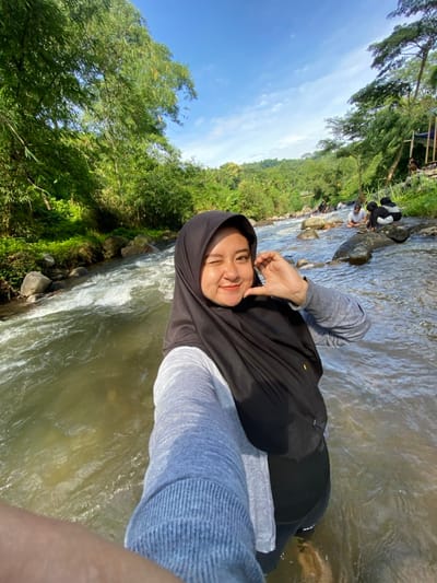 A woman in a black hijab and gray shirt stands in a river. Lush green trees and a blue sky with a few clouds form the backdrop. People are seen along the riverbank in the distance. The water flows around her legs. She is smiling and winking, holding up two fingers in a peace sign. The photo is taken at an angle, making the scene slightly tilted.