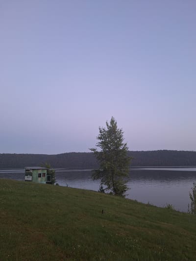 A wide-angle, low-light, outdoor, eye-level shot captures a serene natural landscape. The scene is dominated by a body of water, likely a lake or river, reflecting the soft hues of a twilight sky. The sky itself is a gradient of pale blue and lavender, suggesting either dawn or dusk.
In the foreground, a grassy slope slopes down towards the water. The grass is green and appears somewhat unkempt, with scattered white seed heads visible. A small, weathered, light green structure, resembling a small cabin or shed with a peaked roof and windows, is situated on the left side of the slope. Next to this structure, a young tree with sparse foliage is visible.
Further back, a larger tree with a fuller canopy stands to the right of the cabin. Its leaves are a deep green, providing a contrast to the lighter tones of the grass and sky. The middle ground is occupied by the body of water, its surface rippled by a gentle breeze.
Beyond the water, a dense line of dark green trees forms a treeline along a distant, undulating hillside. The foliage of these trees is so thick that individual trees are indistinguishable, creating a dark, textured mass against the lighter sky. The overall impression is one of quiet solitude and natural beauty, possibly in a rural or park-like setting. The location is not specified in the image or the provided hints.