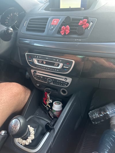 The image shows the interior of a car, likely a hatchback, parked on a street. The dashboard features a digital display showing "20°" on both sides, indicating the air conditioning settings. There are ventilation grilles, a hazard light button, and decorative red bows attached to one of the grilles. A manual gear stick with a fabric scrunchie on its base is visible, along with a key holder with red charms. Below the gear stick, a "START STOP" button and a power outlet are present. On the passenger side, a plastic water bottle is placed on the floor mat. The image is taken from the driver's perspective, showing the dashboard, center console, and part of the passenger seat. The car appears to be parked in a sunny outdoor environment. Based on the provided context, the location is identified as Byala Slatina, Bulgaria.