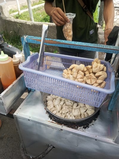 An outdoor scene in Naung Bo Oe, Myanmar, depicts a street food vendor's stall. A person, visible from the chest down, wears a dark green t-shirt and holds a small clear plastic bag containing several skewered, fried food items in their left hand. The stall itself features a metal countertop with a blue wooden frame structure above it. Hanging from the frame are large silver tongs. Resting on the frame is a light purple perforated plastic basket filled with irregularly shaped, lightly browned, fried food pieces. Below this basket, a large metal pan is embedded into the countertop, containing a substantial quantity of similar, paler-colored, irregularly shaped food items. Dark, oily residue stains the countertop around the pan. To the left, two plastic squeeze bottles, one orange-brown with a light green cap and a smaller clear one with a white cap, stand on the counter. A black corrugated hose extends from the bottom left of the metal cart. The background shows a concrete wall, green foliage, and a blurred vehicle on the far left, indicative of an urban or roadside setting.