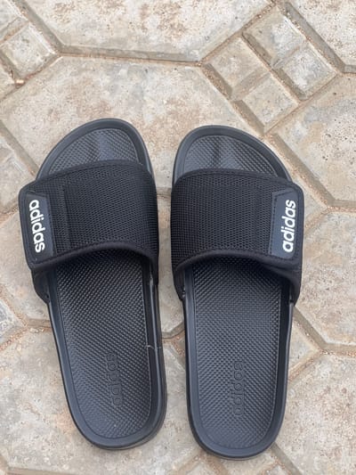 A pair of black Adidas slide sandals are positioned side-by-side on a textured, light-colored stone patio. The sandals are oriented with the toes pointing towards the top of the frame. The left sandal is slightly angled inwards, and the right sandal is positioned to its right and slightly further up. Both sandals feature a black textured footbed with the Adidas logo embossed vertically on the inside edge. The upper strap of each sandal is made of a black mesh material with a black band across it, also bearing the white Adidas logo printed vertically. The sandals are placed on a patio constructed with rectangular and square interlocking stones, some of which are slightly raised. The ground appears to be outdoors in Yali, Nigeria.