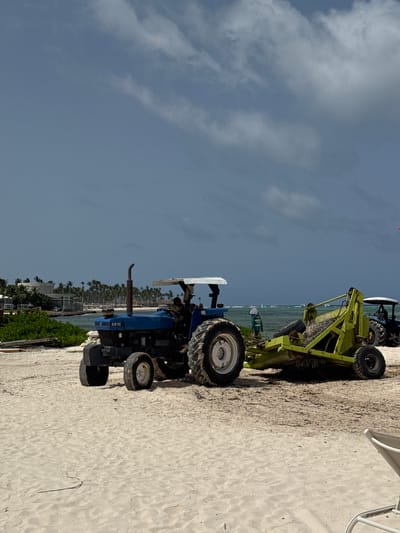 The image shows two tractors on a sandy beach, with the ocean in the background. One tractor is blue and appears to be a utility tractor, with the number "6610" visible on its side. The other tractor is lime green and is attached to a piece of equipment that looks like a tiller or cultivator.
The scene appears to be a beach resort or a similar coastal area, with palm trees lining the shore and buildings visible in the distance. The sky is cloudy, suggesting it might be a partly sunny day. The sand is light in color and appears well-groomed.
The tractors are positioned on the beach, likely for maintenance or landscaping purposes. The presence of the tiller suggests that the area is being prepared or maintained. No people are clearly visible in the immediate vicinity of the tractors.
The overall impression is of a functional operation taking place in a recreational setting. The image captures a blend of industrial machinery with a tropical beach environment. The lighting and cloud cover suggest it is daytime. The location is Higüey, Dominican Republic.