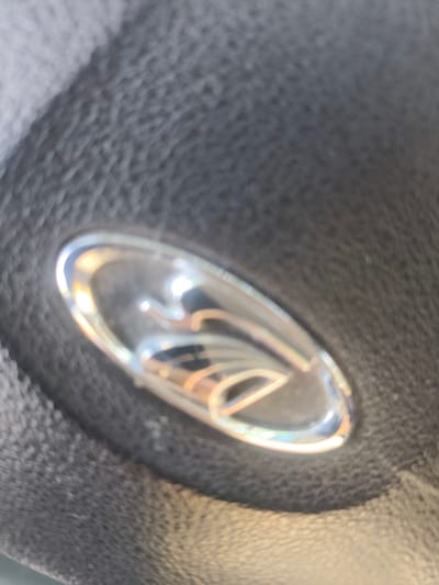 The image shows a close-up, slightly out-of-focus view of a chrome emblem with a reflective surface on a textured, dark grey material, likely leather, consistent with a car's steering wheel. The emblem appears to be a stylized shape with curved lines, possibly a logo. The lighting creates bright reflections and highlights on the emblem. There are no people or any identifiable city landmarks present. The setting is domestic, within a vehicle.