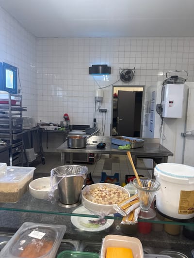 The image shows a commercial kitchen in Rivesaltes, France. The kitchen is equipped with stainless steel countertops, tiled walls, and various cooking appliances. On the left, a stainless steel rack holds trays and stacks of plates. In the center, a stainless steel table has a pot of food and a baking dish. To the right, a large white container and a glass with breadsticks are visible. The overall impression is of a functional, working kitchen environment.