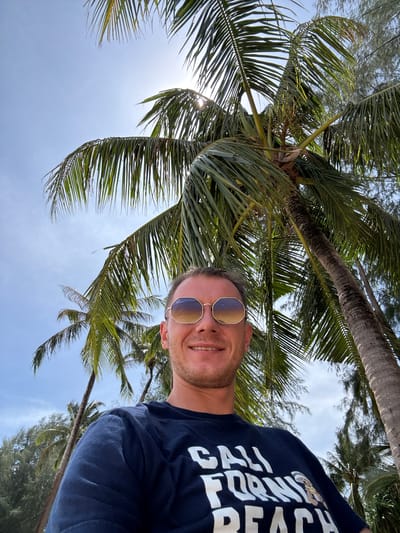 A man wearing sunglasses and a dark blue t-shirt with white lettering stands outdoors under palm trees. The man is smiling and looking at the camera. The t-shirt has the words "CALI FORNI BEACH" printed on it, along with an image of a bear wearing a sailor's hat. The background consists of palm trees and a bright blue sky. The location is Ban Khuk Khak, Thailand.