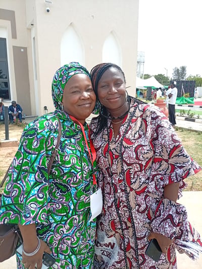 Two women, dressed in vibrant traditional African print attire, pose together and smile at the camera outdoors. The woman on the left wears a green, blue, and pink patterned headscarf and a matching dress. She has an ID lanyard with a white card around her neck, a brown shoulder bag, and is holding a black smartphone with a silver camera module. The woman on the right wears a red, brown, and white patterned dress, with her hair in braids and sunglasses perched on her head. She also has an ID lanyard, part of which is visible.

They appear to be at an outdoor event during bright daylight, suggested by the clear skies and shadows. In the background, there is a light-colored building with arched windows or doorways, possibly a mosque or public building, equipped with a security camera and a small solar panel. Several other people are visible in the distance, some seated and some standing near white and green tents, indicating a gathering or event. Sound equipment, possibly speakers or screens, can also be seen. The setting includes manicured green spaces and paved areas, suggesting an organized venue.

Visible text on the woman on the left's lanyard or a document held in her hand includes "KADUNA," "2026," and "PRINTS NEW CHICA." On the woman on the right's lanyard, "KADUNA STATE CHAPTER," and "2026" are partially legible, along with an acronym that could be "RW" or "RNW." Both women exhibit warm smiles, conveying a friendly and joyful mood. The attire and general setting are consistent with the provided location context of Unguwan Sanusi, Nigeria.