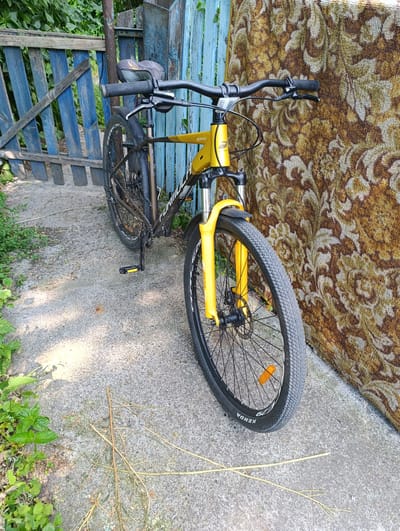 This media file captures a yellow and black bicycle parked on a concrete path next to a blue wooden fence and a patterned fabric tapestry. The bicycle is the primary object, featuring a suspension fork and knobby tires, suggesting it's a mountain or hybrid bike. The frame has visible branding, with "FORMULA" written vertically on the down tube and some unreadable text in yellow. The front tire has "KENDA" printed on it. The scene appears to be outdoors, with greenery and foliage visible in the background and to the left of the path, indicating a natural or garden setting. The sunlight suggests it's daytime, possibly mid-morning or afternoon, casting shadows on the concrete. The blue fence, with its weathered appearance and a slightly damaged section, combined with the patterned tapestry leaning against it, creates a somewhat rustic and perhaps domestic environment. There are no people visible in the image, and the overall mood is still and tranquil. The location context provided is Fastiv, Ukraine.