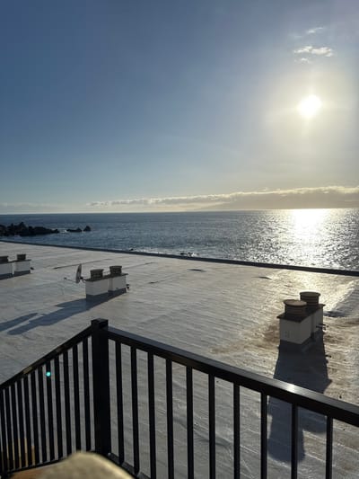 The image displays a coastal scene from an elevated perspective in Santiago del Teide, Spain, under clear, sunny conditions. In the foreground, a black metal railing with vertical balusters partially obstructs a light grey flat rooftop. On this rooftop, three white, square-based ventilation shafts with brown circular caps are visible; one shaft on the left features an attached small, light-colored satellite dish. These structures cast elongated shadows towards the viewer. The midground reveals the Atlantic Ocean, with waves gently breaking against a rocky shoreline on the left. The sun, a brilliant white glare, is positioned in the upper right quadrant, creating a strong reflective path across the water's surface. The background consists of a predominantly clear blue sky with a distinct, horizontally stretched band of low-lying clouds along the distant horizon, partially obscuring a landmass. No people are present in the scene.