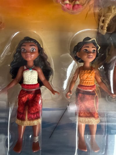 The image shows two Moana dolls in their original packaging. The dolls are oriented vertically, with the heads pointing towards the top of the frame. Both dolls have dark hair and brown eyes. The upper doll is wearing an orange top and a reddish-orange skirt. The lower doll is wearing a white and red patterned top and a reddish-orange skirt. Both skirts have gold trim. The dolls appear to be made of plastic. The background is a light brown and blue gradient, with some blurry elements that suggest they are inside a box or display. There is no visible text in the image. The lighting is bright, suggesting it was taken in daylight. The overall impression is of a product for sale, likely in a store or as a gift. The context of Chociule, Poland is noted, but there are no visual cues in the image to confirm this location.
