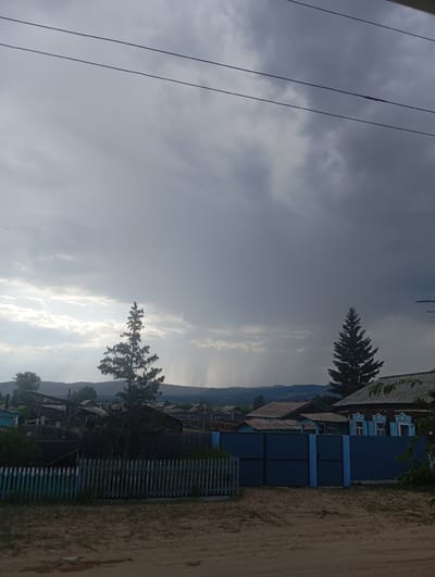 This is a landscape photograph taken in Novodesyatnikovo, Russia. The scene depicts a rural village under a cloudy sky, with visible rain shafts in the distance. The foreground is a dirt path and a low, light blue and white picket fence with overgrown grass. Behind the fence, a dark blue metal gate stands open, leading into a yard with more vegetation.
Several wooden houses with traditional Russian architectural details, such as carved window surrounds, are visible behind the blue gate and further into the village. Some of these houses have corrugated metal or shingled roofs. There are also fences surrounding the properties, made of various materials including wood and metal. Two tall evergreen trees stand prominently in the mid-ground, one to the left of a large blue gate and another to the right, silhouetted against the sky. The background shows rolling hills and mountains, suggesting a natural landscape surrounding the settlement. Overhead, two dark electrical wires stretch across the frame, adding a man-made element to the natural scenery. The overall atmosphere is overcast, with patches of sunlight breaking through the clouds and illuminating the distant rain.