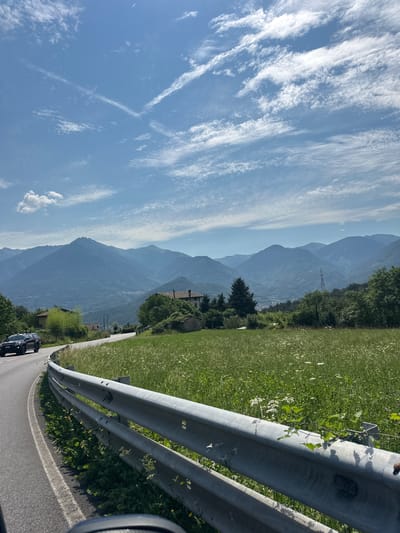 The image shows a road winding through a mountainous landscape in Ossimo Inferiore, Italy. The road is paved and bordered by a metal guardrail. To the right of the road is a field of green grass dotted with small white flowers. In the distance, several layers of mountains are visible, with the furthest mountains appearing hazy blue. The sky is a clear blue with scattered white clouds. A dark-colored SUV with "CARABINIERI" written on its side is visible on the road, driving away from the viewer. A building with a red tile roof and a small stone structure are nestled among trees on a hillside.