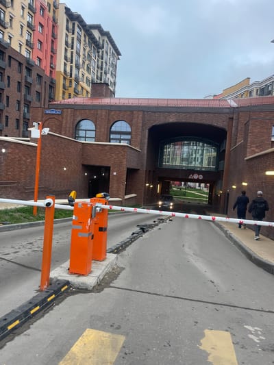 A ground-level, slightly tilted, outdoor shot depicts a barrier at the entrance to an arched brick structure, likely a parking garage, in Razvilka, Russia. The building on the left has multiple sections in different colors, including red, yellow, and brown. The address sign "РИМСКИЙ 15к1" (Rimskiy 15k1) is visible on the side of this building. The barrier has a white and red striped arm. Two orange posts hold up the barrier's mechanism, with yellow warning lights on top. A black vehicle with its headlights on is driving toward the camera in the distance, passing through the archway. Pedestrians are seen walking on the walkway behind the vehicle. The sky is overcast. The pavement in front of the barrier has yellow markings.