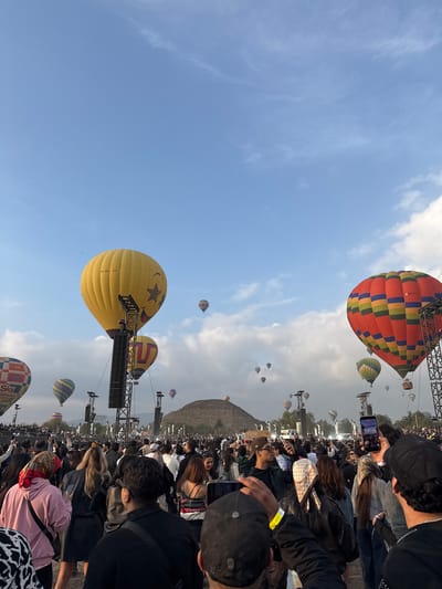 The image captures a vibrant scene from San Francisco Mazapa, Mexico, filled with a crowd of people watching hot air balloons. Several hot air balloons of different colors and designs are visible in the sky, including a yellow one with a smiley face. The crowd is dense, with people holding up phones to capture the spectacle. Structures such as metal frameworks are interspersed among the balloons. The sky is partly cloudy, and some mountain ridges are visible in the background.