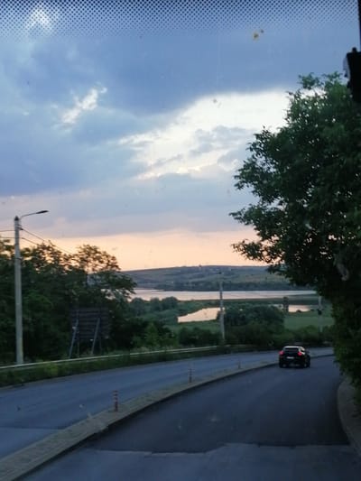 A black SUV is driving on a paved road. The vehicle is seen from the rear, and its brake lights are illuminated. The road curves to the right, and there is a sidewalk on the left side of the road. The sidewalk is bordered by a grassy area and a metal guardrail. Beyond the guardrail, there are trees and a body of water. The sky is cloudy with a sunset in the background. The scene appears to be in a rural or suburban area. The presence of a street light suggests it is a developed area, possibly on the outskirts of Fălticeni, Romania, where such roads and landscapes are common.