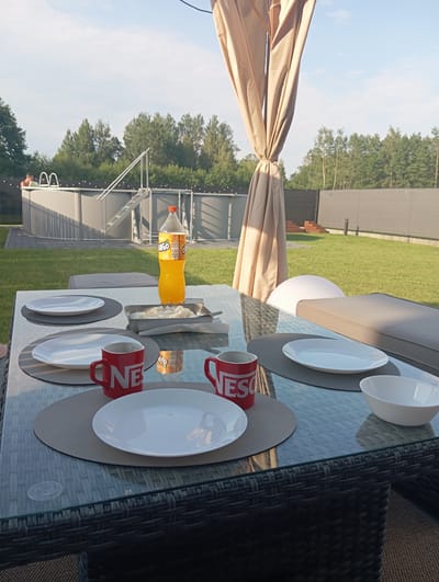 A bright, outdoor scene is captured from a low angle, focusing on a glass-topped dining table set for a meal. The table is made of rattan and is positioned on a patio or deck. On the table, there are several white plates, two red mugs with the "Nescafé" logo, a metal dish containing what appears to be mashed potatoes, and a bottle of orange soda. A beige canopy is visible in the background, casting shadows. Beyond the table, a green lawn stretches out, leading to a fenced area with a swimming pool and a person standing near its edge. Trees form a dense line along the horizon under a clear blue sky with scattered clouds. The overall setting suggests a leisurely meal outdoors, likely in a suburban or rural area of Dybów Kolonia, Poland.