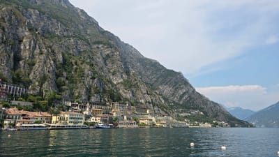 A scenic wide shot captures a sun-drenched view of Limone sul Garda, Italy, from the tranquil waters of Lake Garda. The town, nestled at the base of a towering, rugged mountain, is characterized by its cluster of colorful buildings with terracotta roofs. These structures cascade down the mountainside, some appearing to be hotels or residences, with balconies and awnings visible. The lake itself is calm and dark blue, with gentle ripples reflecting the light. Several white buoys bob on the water's surface. A small blue and white boat is docked near the shore, alongside other vessels and a pier lined with tables and chairs, suggesting a lively waterfront. The steep mountain dominates the background, its rocky slopes interspersed with patches of green vegetation. The sky is a pale blue with scattered, wispy clouds, hinting at fair weather. In the distance, the hazy outlines of more mountains can be seen, adding depth to the breathtaking landscape.