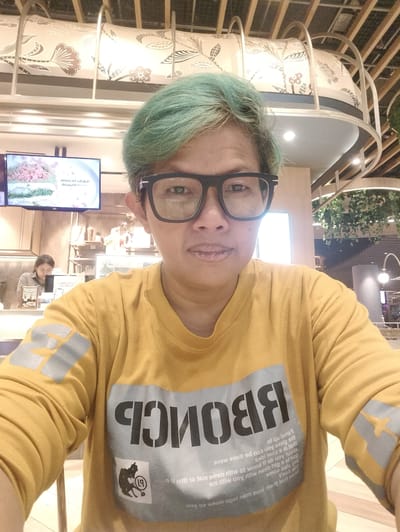 In Gunung Putri, Indonesia, a person with turquoise hair and black-rimmed glasses is pictured in a selfie. They are wearing a mustard yellow long-sleeved t-shirt with large, dark grey lettering on the front that reads "BRONCE" in reverse. The t-shirt also has a smaller graphic of a person with the number 19 inside a circle. The background appears to be a cafe or restaurant interior, with wooden beams and decorative elements visible on the ceiling. To the left, a television screen displays images of food, and a person is seen working behind a counter. The lighting suggests it is daytime, likely indoors.