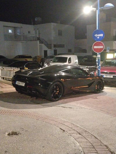 A black McLaren supercar, with license plate NNN 777, is parked on a paved area with brick accents, likely in Ayia Napa, Cyprus. The car is sleek and low to the ground, with distinctive orange accents on its side. In the background, several other vehicles are visible, including a white van and a red sedan with license plate EZB 632. A blue lamppost with two traffic signs – a blue sign with a white arrow pointing left and a red and white "no entry" sign – stands to the right of the McLaren. The scene appears to be a street or parking lot at night, with artificial lighting illuminating the area. Buildings with windows are visible in the background, suggesting an urban or commercial setting.