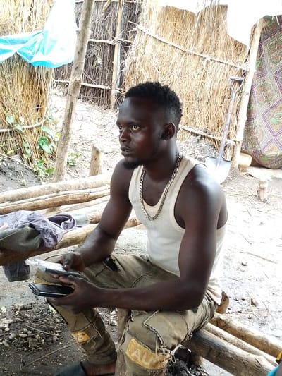 A dark-skinned man with short, dark hair and a short beard sits on a wooden bench, holding two black cell phones. He wears a white tank top and tan, distressed pants with yellow patches. He is seated outdoors, in what appears to be a rural or informal setting, possibly in Kajola, Nigeria. Behind him is a structure made of reeds and bamboo, with a blue tarp draped over it on the left. A shovel leans against the reed structure on the right, and a colorful patterned fabric hangs to the far right. The ground is dirt and scattered with small stones and debris. The man's posture and grip on the phones suggest he is examining or using them.