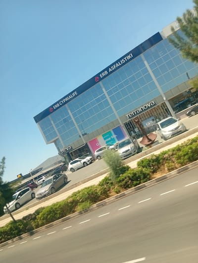 The image captures a bright, sunny daytime scene in Paralimni, Cyprus, focusing on a large, modern commercial building with a striking reflective glass facade. The building prominently features the signs "▲ ERB CYPRIALIFE" and "▲ ERB ASFALISTIKI" on its upper sections, indicating insurance or financial services. On the ground floor, a "Terranova" fashion store is visible, with a partially obscured slogan that reads "Co your life." nearby. A large, rusty decorative anchor sculpture stands in front of the Terranova entrance.
Numerous cars of various models and colors are parked in front of and around the building, including a multi-level parking structure with a canopy visible further back, suggesting a busy commercial area. A paved road with white dashed lane markings runs across the foreground, separated from the parking areas by a curb and a strip of green bushes and small trees. A distant yellow "Lidl" logo is visible on another building. The clear blue sky and bright sunlight indicate fair weather. The scene depicts a typical commercial street or retail park, with vehicles parked, implying people are shopping or working.