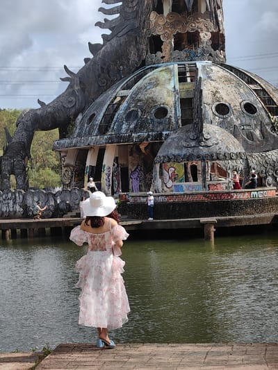 An outdoor scene in Kim Long Ward, Vietnam, depicts a prominent derelict dragon-themed structure overlooking a body of water. In the foreground, a person stands on a reddish-brown paved surface, viewed from behind, facing the water and the structure. This individual wears a wide-brimmed white hat, a white and pink floral ruffled off-the-shoulder dress, and light blue open-toed block heels, holding a light pink handbag.
The midground features a calm body of greenish-brown water. On the far side of the water, a concrete platform forms the base of the large structure. Several individuals are present on this platform: one person in light clothing crouches on the far left; two people in white tops and dark pants stand upright in the center and center-right, respectively; and a group of three to four individuals, including one in a red garment and straw hat, are situated further right near the structure's base.
The dominant background subject is a large, weathered structure resembling a dark grey dragon coiled around a dilapidated domed building. The dragon's textured body and head rise prominently. The domed building beneath is heavily discolored with patches of grey, green, and blue, featuring numerous broken or empty window openings, some revealing internal stairs. Extensive multi-colored graffiti covers the lower walls and columns of the building and the concrete platform. Dense green foliage and trees are visible on a hill behind the structure to the left. The sky overhead is overcast with grey clouds.