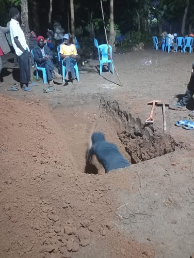 The image depicts an outdoor nighttime scene in Butali Chegulo ward, Kenya, centered around the excavation of a deep pit in reddish-brown earth. In the foreground, one individual, clad in dark blue clothing, is partially submerged within the rectangular pit, actively engaged in digging, with their torso visible. A pile of freshly excavated earth surrounds the pit. Adjacent to the pit, a small shovel with an orange handle and a pickaxe lie on the ground.

To the left and in the background, approximately ten adult males are present. One man stands in the immediate left foreground, wearing a white short-sleeved shirt and dark trousers, with blue sandals on the ground nearby. Further back, two men are seated on light blue plastic chairs; one wears a red cap and a dark jacket, while the other wears a yellow short-sleeved shirt and a light blue cap. Additional men are seated on similar blue chairs in the mid-ground and background.

The ground is predominantly bare, reddish-brown soil, appearing somewhat moist or reflective in patches. The area is illuminated by an unseen light source, casting shadows. Trees and dense foliage form the background, indicating a rural or semi-rural setting. Several pairs of sandals are visible on the ground near the seated individuals, and a long wooden stick leans against an empty blue chair in the mid-ground.