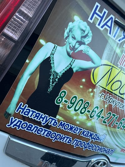 The image features a sign partially visible above a car's taillight. The sign displays a photograph of Marilyn Monroe, a phone number (8-908-64-271-64), and text in Russian. The text includes the name "Natalya" and the city "Irkutsk." The overall aesthetic appears dated and stylized, potentially advertising a service or business. The image's angle suggests it was taken in close proximity to the subject. The presence of the phone number, and the phrase "Natalya," along with the city Irkutsk, suggests that the location in Sayansk, Russia, may be related to the content of the sign.