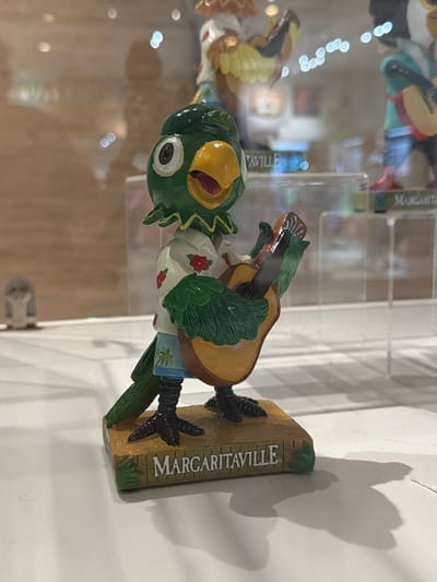 A close-up shot shows a decorative figurine of a green parrot playing a guitar. The parrot is wearing a white shirt with red floral patterns and blue shorts with a palm tree design. It stands on a wooden base with the word "MARGARITAVILLE" inscribed on it. The figurine is placed on a white surface, possibly a display shelf, with a blurred background that suggests an indoor environment, possibly a store or museum, with reflections of lights and other objects visible. The lighting is soft, casting a shadow of the figurine to the right. The image quality is slightly blurry, especially in the background.