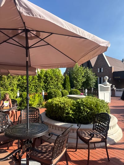An outdoor seating area is depicted with a large, light-colored umbrella providing shade. Under the umbrella, a round, ornate metal table and chairs are arranged on a brick patio. A woman is seated at one of the tables, using a laptop. The background features a well-maintained garden with manicured hedges and trees, a stone fountain, and a white decorative fence with posts. In the distance, a brick building with a tiled roof is visible, bearing a sign with Cyrillic lettering that reads "ВЯЗОВАЯ" (Vyazovaya). The scene appears to be a cafe or restaurant in Seltso, Russia, on a sunny day.