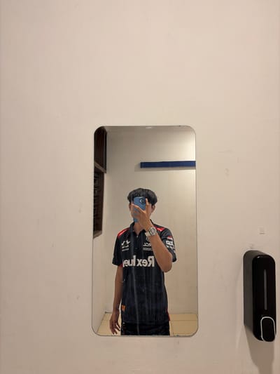 In the reflection of a mirror mounted on a textured, off-white wall, a person is captured taking a selfie with a blue smartphone. The person, facing left, is wearing a black t-shirt with red and white accents and various logos and text. A prominent watch with a silver band is visible on their left wrist. Their right arm is extended, with the hand open. The mirror is rectangular with rounded corners and has some smudges and marks on its surface. Above the mirror, a black soap dispenser with a silver button is mounted on the wall. The background reflected in the mirror shows a portion of a wall with a blue vertical stripe and a vent with metal bars below it. The image appears to have been taken indoors during daylight hours, given the ambient lighting. The scene is simple and focuses on the individual in the mirror. Visible text on the person's shirt includes "REX", "RE," and "EX."