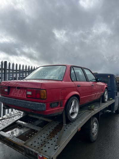 A red car is secured on a tow truck. The scene is an exterior location in Shotts, United Kingdom, under a cloudy sky. A fence is visible in the foreground.