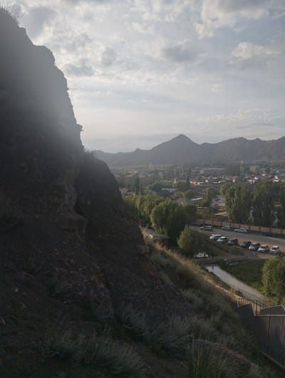 An elevated, wide-angle shot captures a dramatic landscape with a dark, rocky cliff dominating the left foreground. The cliff face is steep and uneven, with sparse vegetation clinging to its surface. Beyond the cliff, a town unfolds, nestled in a valley and surrounded by rolling hills and distant mountains under a cloudy sky. The town features a mix of buildings, including houses and apartment blocks, with trees scattered throughout. A road runs through the town, with several cars parked along its side. A small body of water is visible, crossed by a bridge. Stairs with metal railings descend from the foreground towards the town, suggesting a viewpoint or a path. The overall impression is a blend of natural ruggedness and human settlement. The scene is set in Naryn City, Kyrgyzstan.