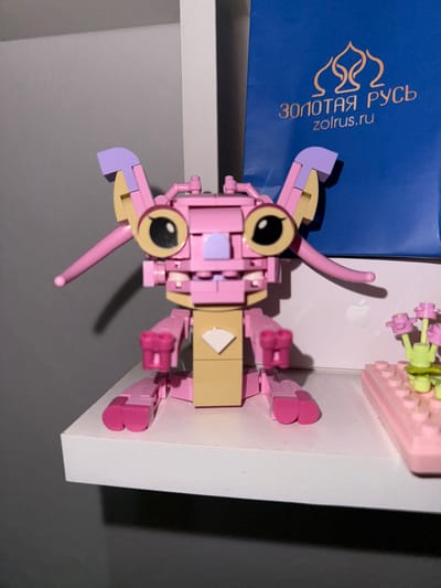 A pink and tan LEGO figure with large black and yellow eyes, purple ears, and thin pink tentacles stands on a white shelf. Behind the figure, a blue bag with gold lettering and an image of a flame-like design is visible. To the right of the LEGO figure, a pink LEGO structure with green and pink floral elements is present. The background consists of a gray wall and a white wall. The scene is indoors.