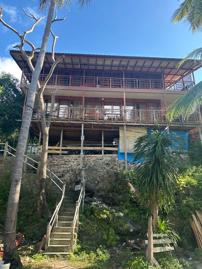 A three-story wooden building with a metal roof is built on a rocky hillside in A R A, Indonesia. The building has balconies on each level, with railings made of metal bars. The ground floor appears to be open, with wooden supports and a blue tarp covering part of the area. Green vegetation, including palm trees and bushes, surrounds the building. A concrete staircase with white railings leads up the side of the hill to the building. There is some debris and trash visible on the ground around the staircase and the base of the building. The sky is clear and blue.