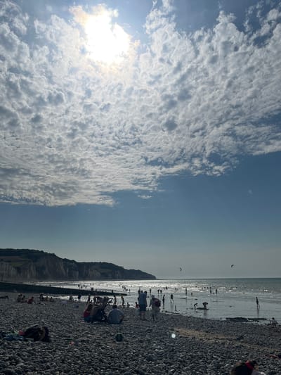 This media file depicts a crowded beach scene on a sunny day with some clouds. The image is oriented vertically, with the sky occupying the left half and the beach and sea the right half. The sky is filled with a dramatic display of white and gray clouds, with the sun shining brightly and casting a warm glow.

On the right side of the image, a pebbly beach stretches along the coastline, with numerous people scattered across it and in the shallow water. The sea is calm and reflects the bright sky, creating a shimmering effect. In the distance, a cliff face rises from the land, adding a dramatic backdrop to the scene. Several people are engaged in various beach activities, including sunbathing, walking, and playing in the water. Some appear to be kiteboarding or flying kites in the distance. The atmosphere seems lively and relaxed, indicative of a pleasant summer day.

The presence of people in swimwear and the general ambiance suggest it's likely midday or late afternoon during a warm season. The clear blue sky with scattered clouds indicates fair weather. The cliff formation and the overall landscape are consistent with a coastal setting like Hautot-sur-Mer, France. No visible text is present in the image.